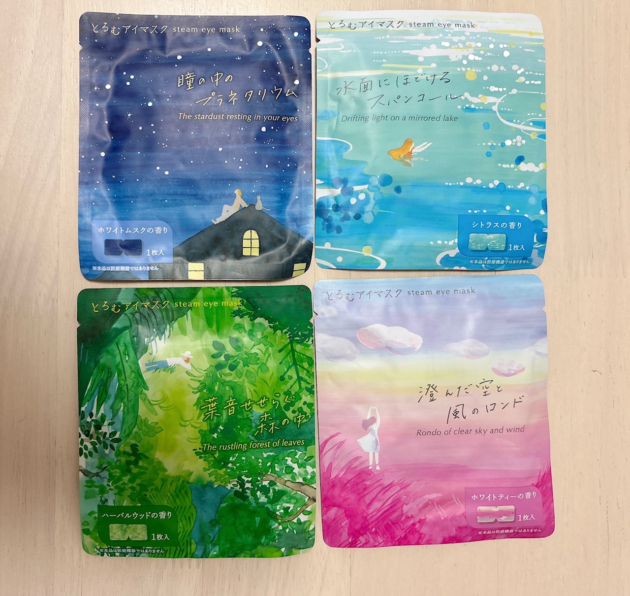 Four colorful steam eye mask sachets laid out on a light wooden surface, each with Japanese text and a whimsical illustration (night sky, lake, forest, and pink sunset). They appear to be assorted eye mask scents or flavors.