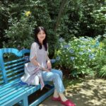 Woman sits on a bright blue garden bench, smiling, with lush greenery and blue hydrangeas nearby.