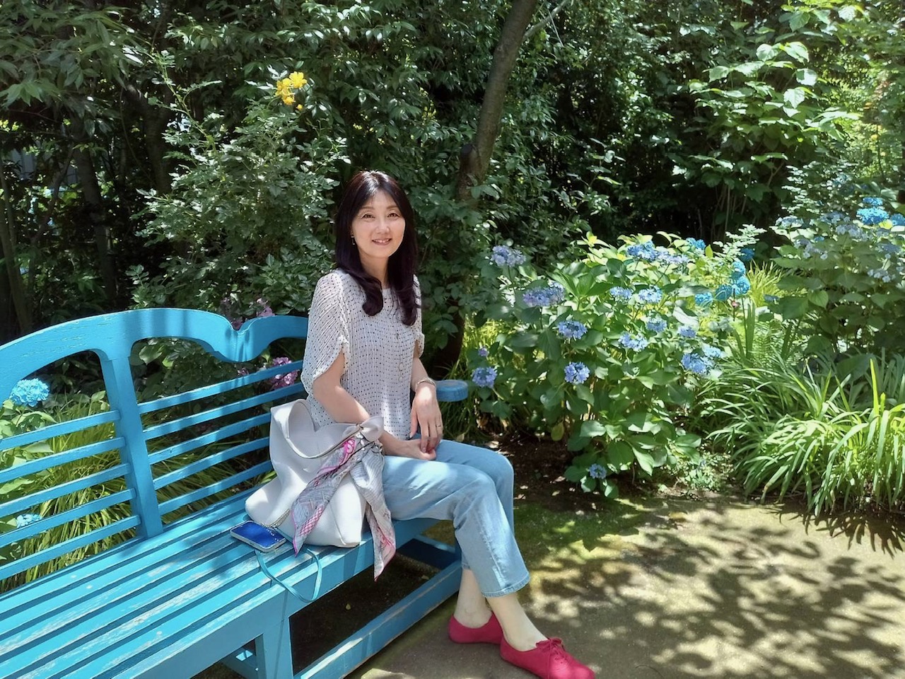 Woman sits on a bright blue garden bench, smiling, with lush greenery and blue hydrangeas nearby.