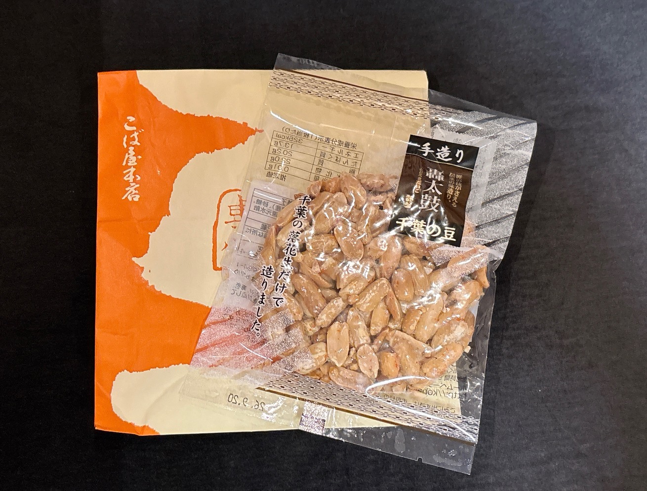 Bag of roasted peanuts in clear plastic package with Japanese text, placed on an orange and cream box/cover, likely a snack