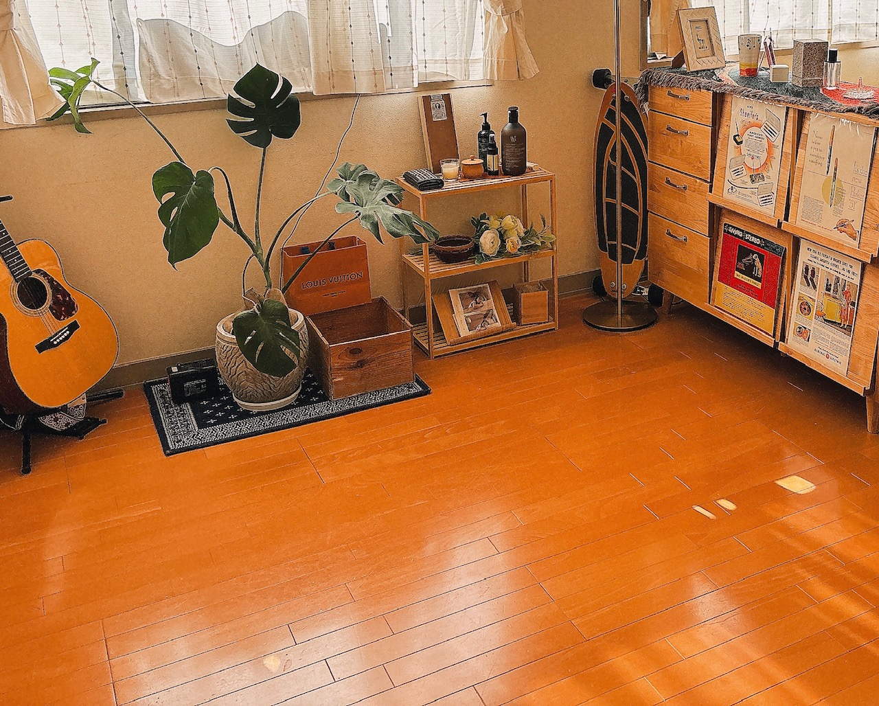 Guitar leaning on a stand beside a large potted monstera, with a small rack of decor, a Louis Vuitton box, and a sunlit wooden floor in a cozy room.
