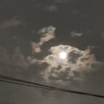 Full moon visible behind scattered clouds in a night sky, with power lines in the foreground.