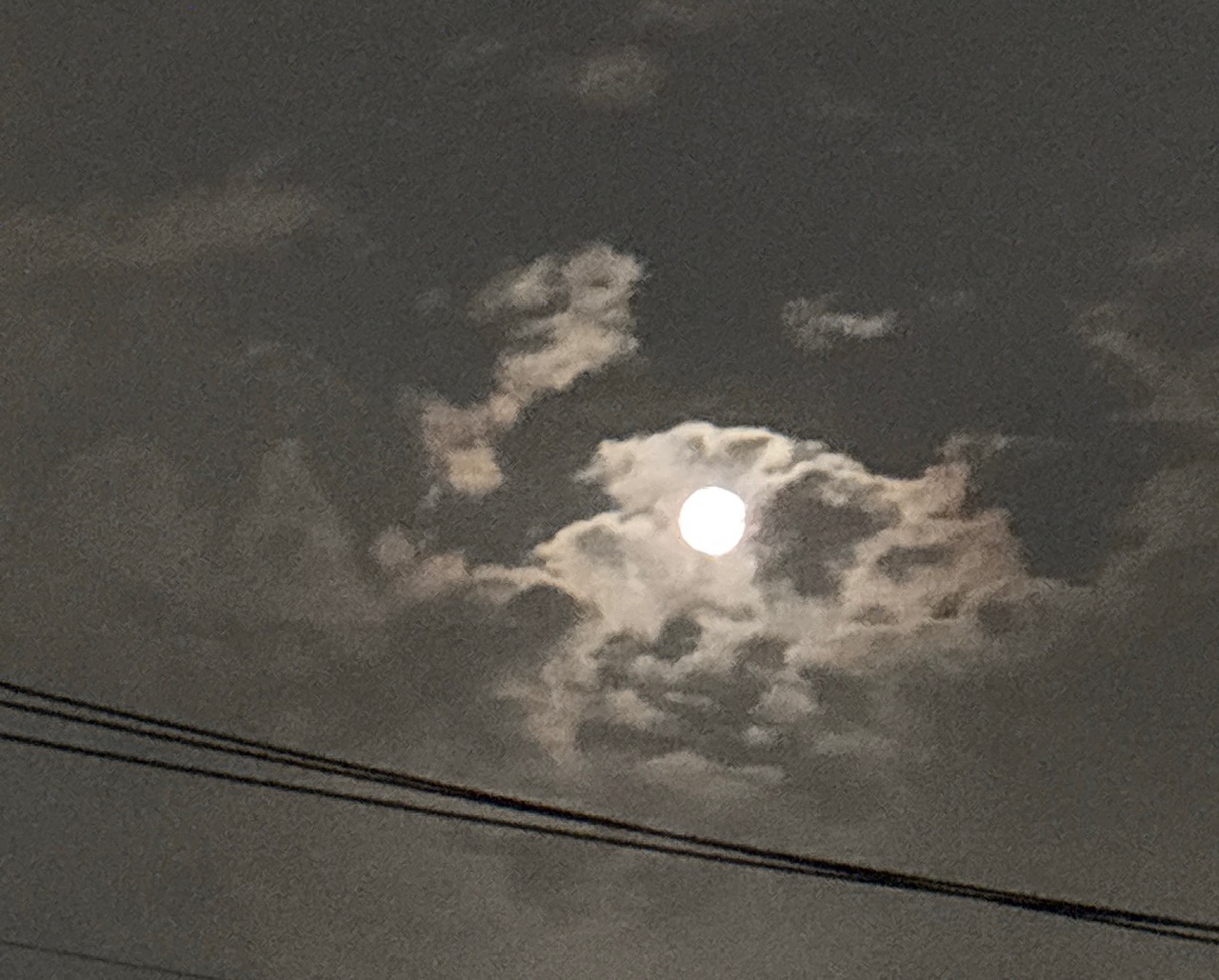 Full moon visible behind scattered clouds in a night sky, with power lines in the foreground.
