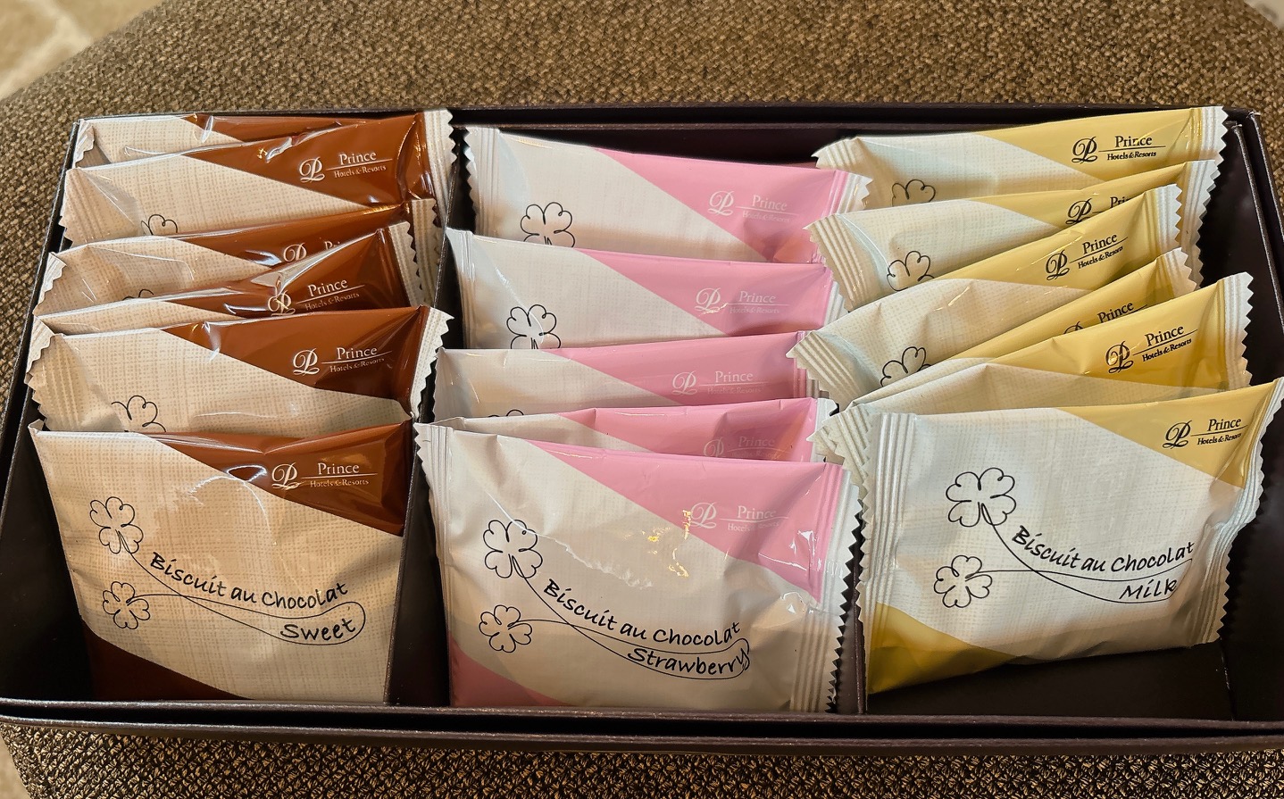 Assorted individually wrapped biscuits in a black box, with brown, pink, and yellow wrappers labeled ‘Biscuit au chocolat’ and ‘Sweet’/‘Strawberry’ flavors (Prince Hotels & Resorts).