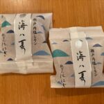 Two individually wrapped Japanese snacks on a wooden surface, paper packaging with silver stripe and blue wave decorations, vertical Japanese text.
