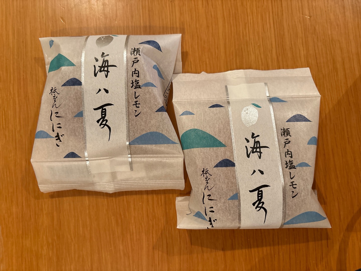Two individually wrapped Japanese snacks on a wooden surface, paper packaging with silver stripe and blue wave decorations, vertical Japanese text.