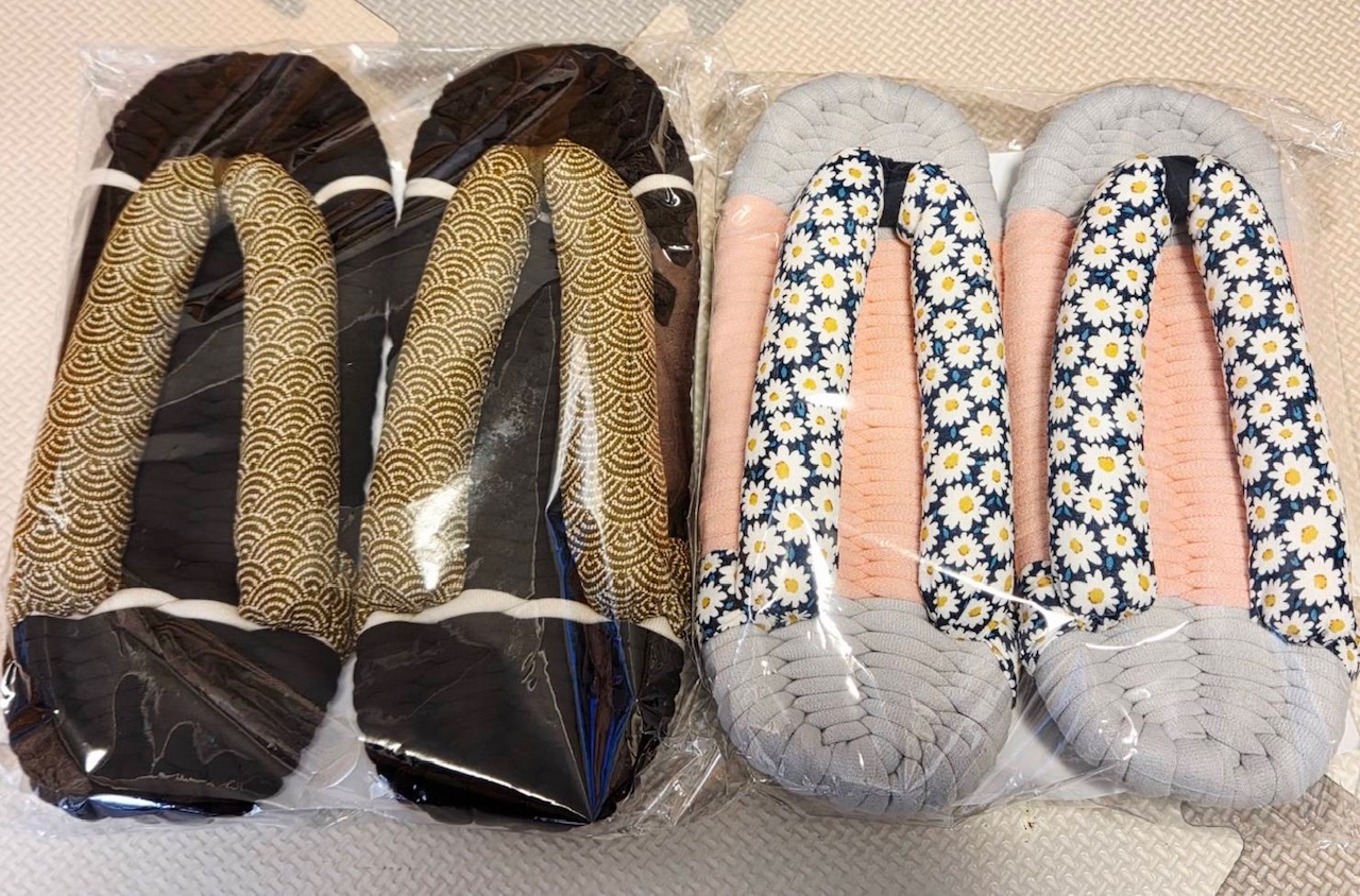 Two packaged pairs of padded sandals: left pair in dark brown with gold wave-pattern straps, right pair in light gray with blue daisy-pattern straps and peach insoles.