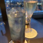 Two glasses on a table with a city skyline and bokeh lights in the background; one tall iced beverage with condensation and ice cubes, and a slender champagne flute nearby.