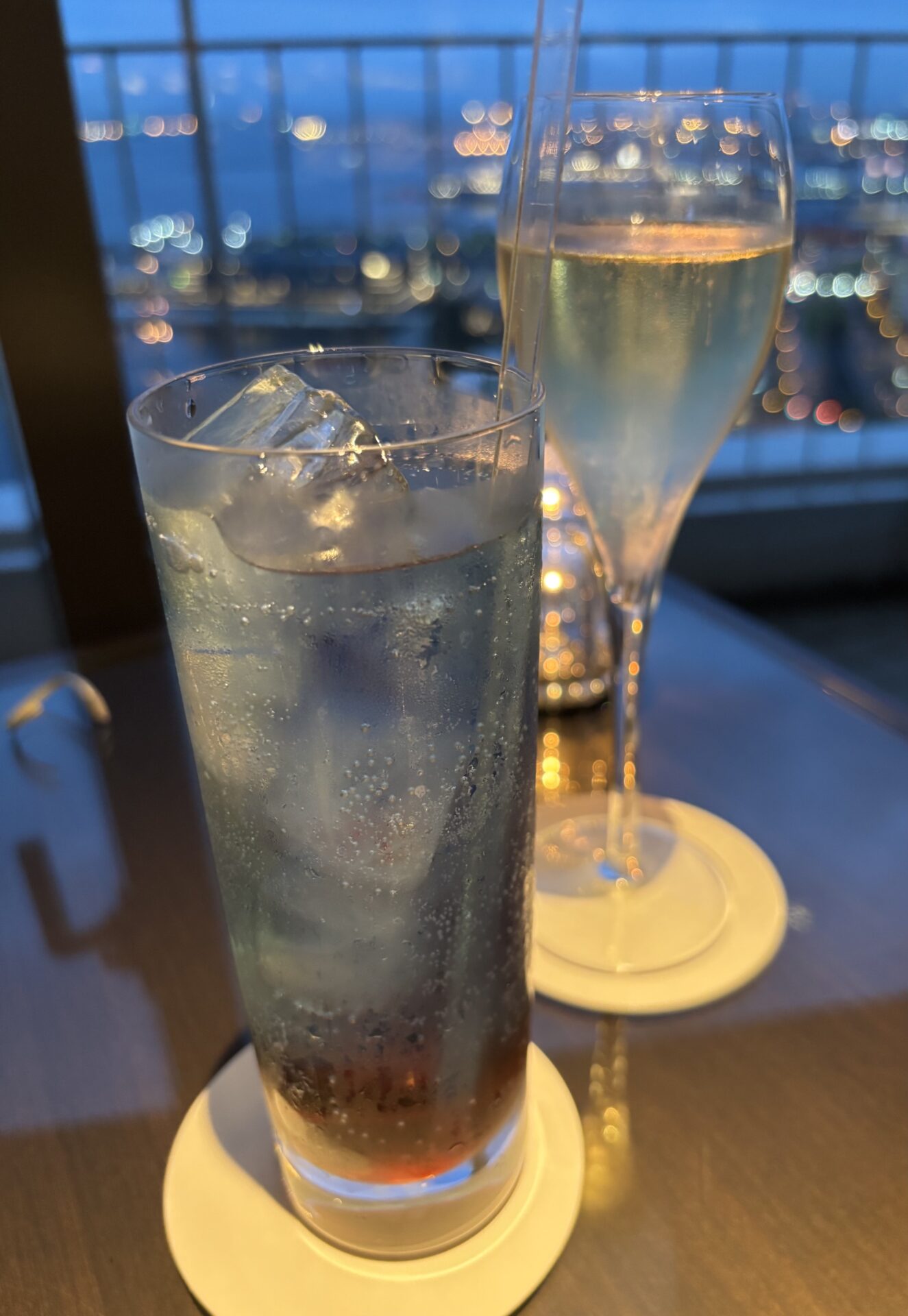 Two glasses on a table with a city skyline and bokeh lights in the background; one tall iced beverage with condensation and ice cubes, and a slender champagne flute nearby.