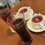 Two white teacups on saucers with tea, beside two tall glasses of iced coffee with red straws on a wooden table.