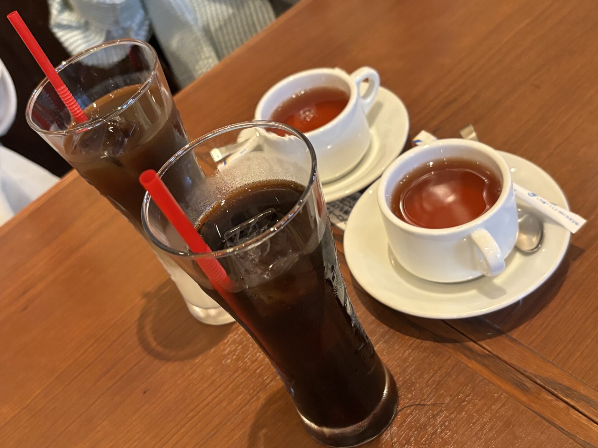 Two white teacups on saucers with tea, beside two tall glasses of iced coffee with red straws on a wooden table.