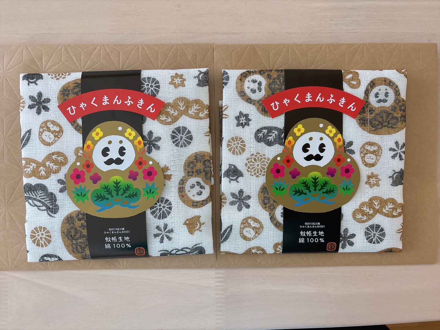 Two fabric gift packs on a brown surface, each with a colorful Daruma motif and a red Japanese banner on white patterned cloth.