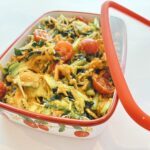 Enamel casserole dish with a colorful veggie pasta salad—tomatoes, greens, and shredded cheese in a yellow sauce, ready to serve.