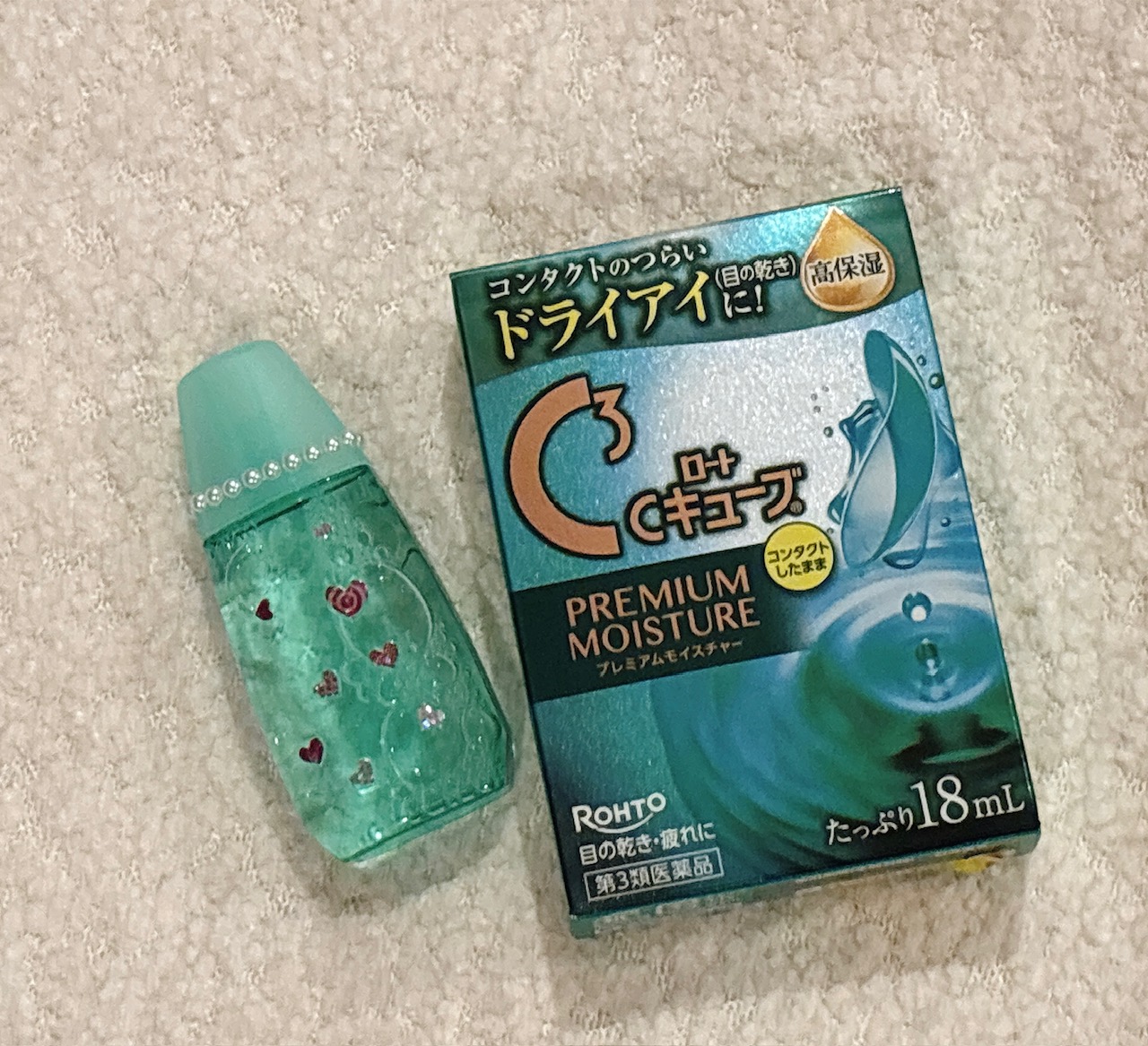 Green contact lens solution bottle with small red hearts next to a teal Rohto Premium Moisture box on a carpeted surface.