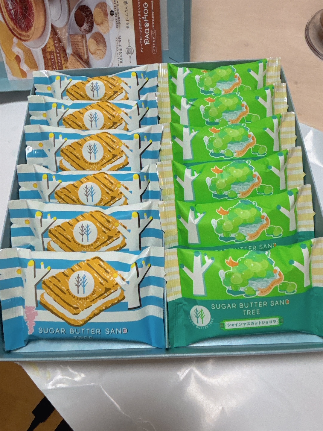 Display box with two stacks of Sugar Butter Sand cookies: blue wrappers on the left featuring a sandwich icon and a white tree, green wrappers on the right with a grape cluster and white trees.