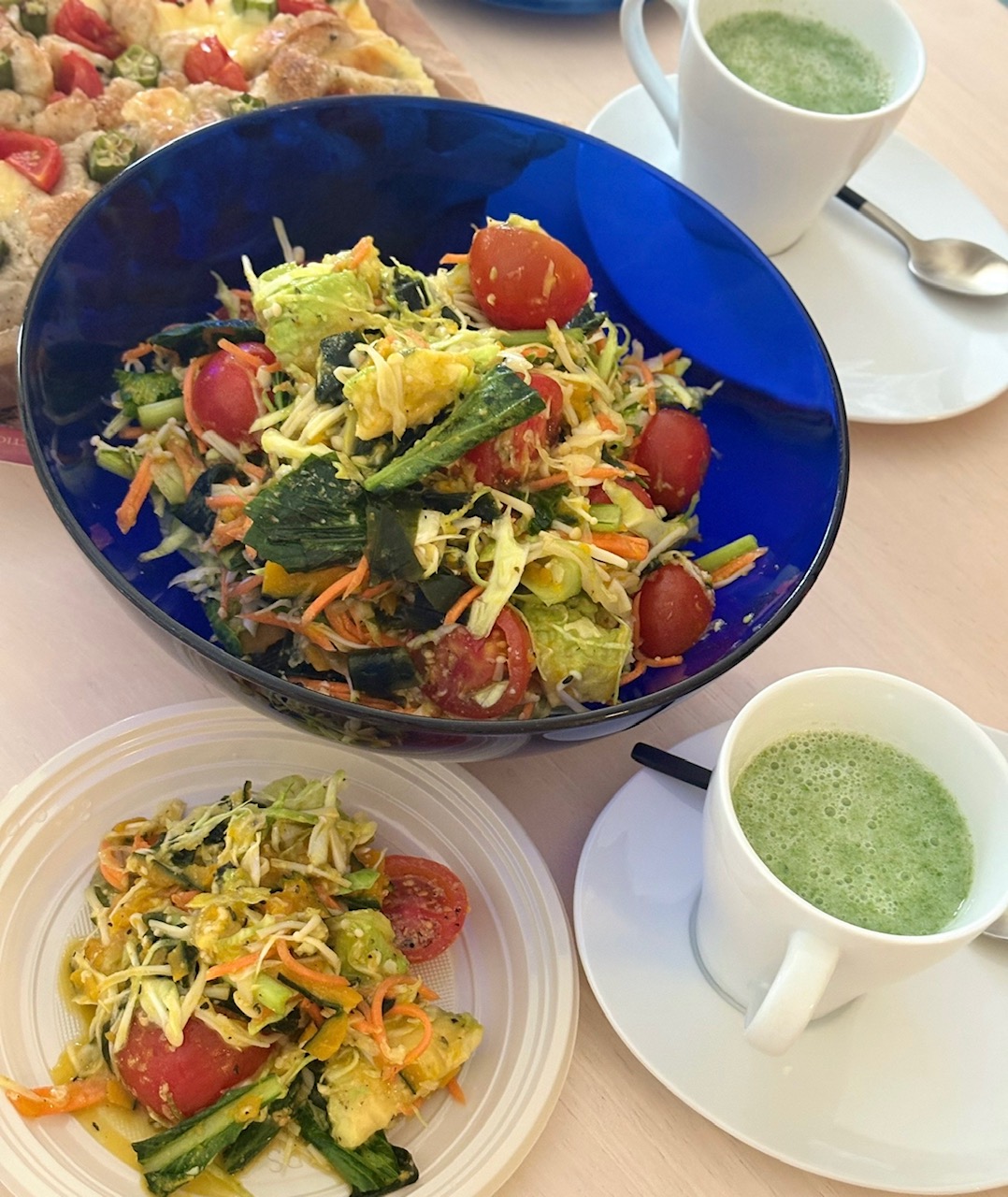 Large blue bowl filled with colorful mixed vegetable salad (lettuce, cherry tomatoes, shredded carrots, cucumber) on a light table, with two cups of green smoothie nearby.